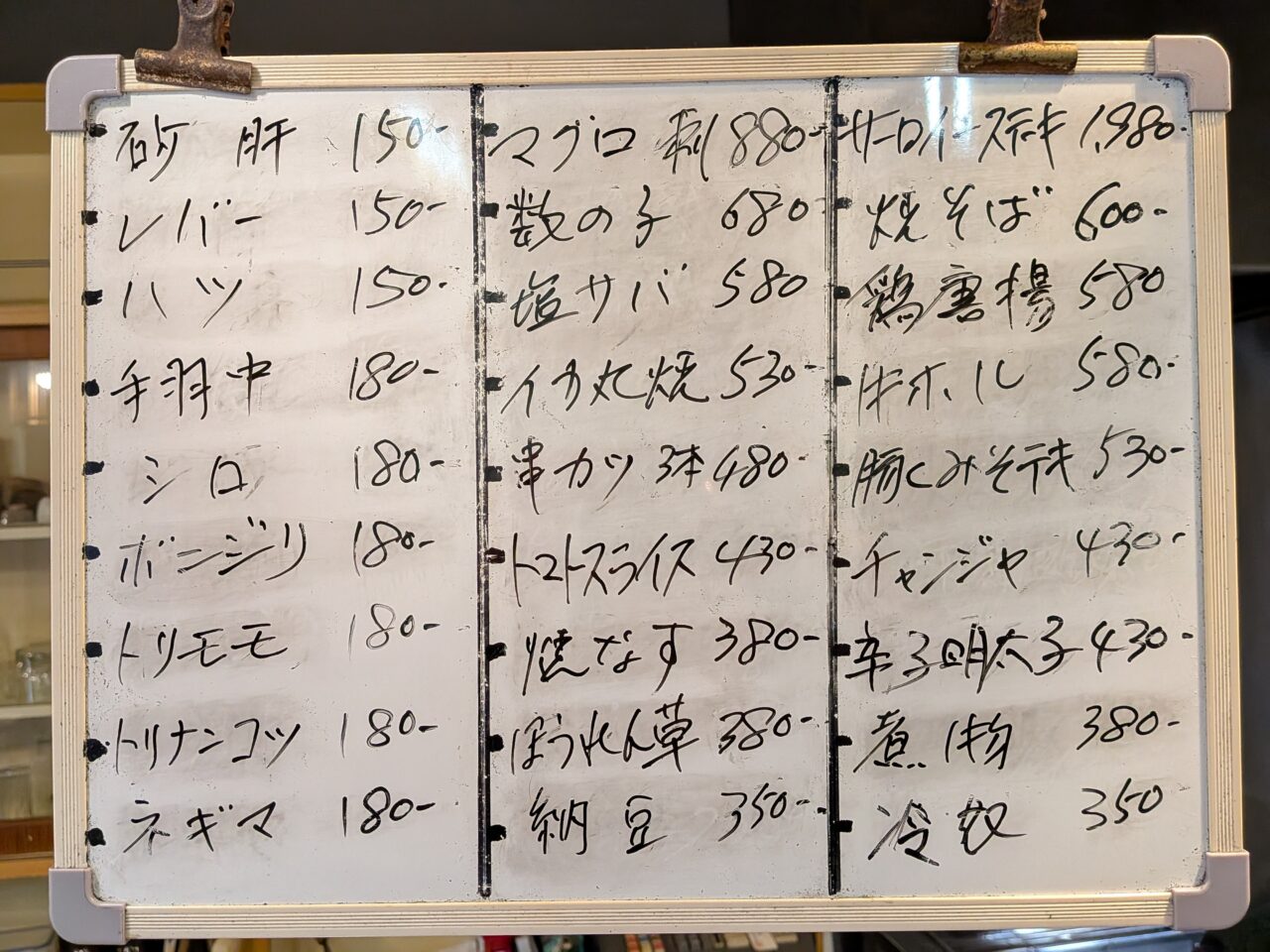 串焼きや一品料理の価格が記された、店内に掲示された手書きのホワイトボードメニュー