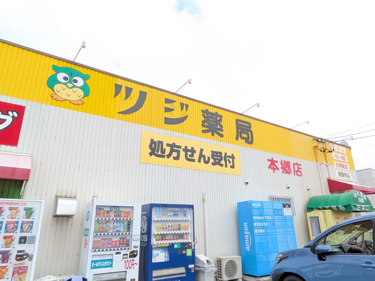 ツジ薬局 本郷店に並ぶ自動販売機と建物外観