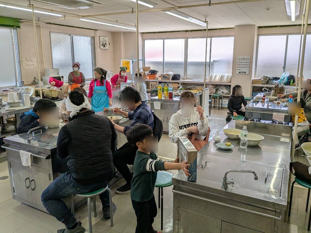 神守小学校区コミュニティセンターの調理室兼会食会場に集まり食事や会話を楽しむ子育て家庭とスタッフの様子